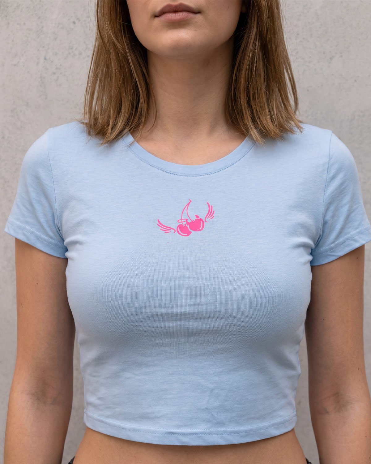 Modelo usando el crop top Cherry celeste de Origo con estampado de cereza alada en rosa neón